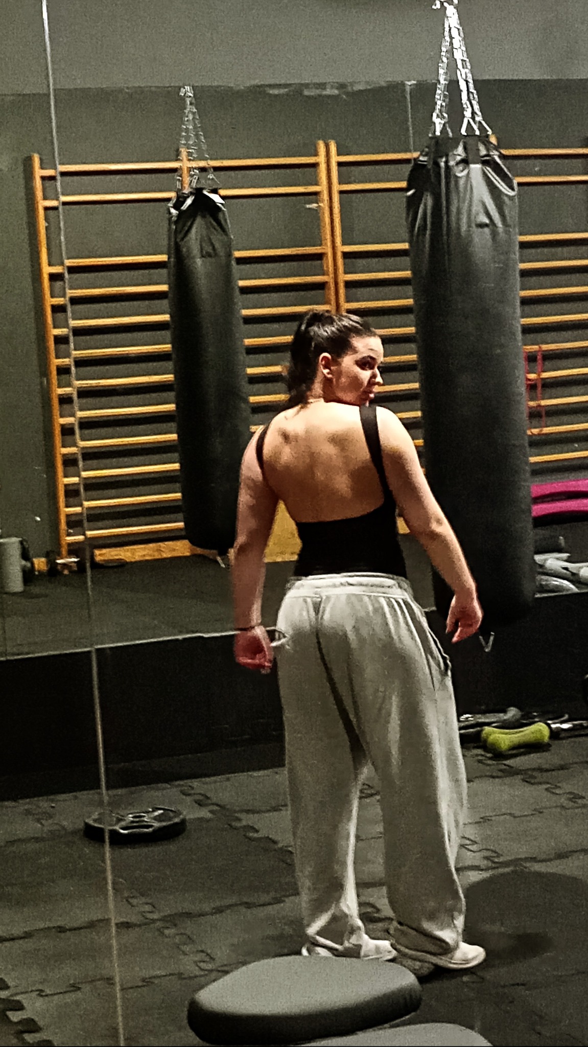 Kobieta w stroju sportowym stoi tyłem w siłowni, widoczne worki treningowe i drabinki gimnastyczne. Oświetlenie stonowane, akcent na sylwetkę.