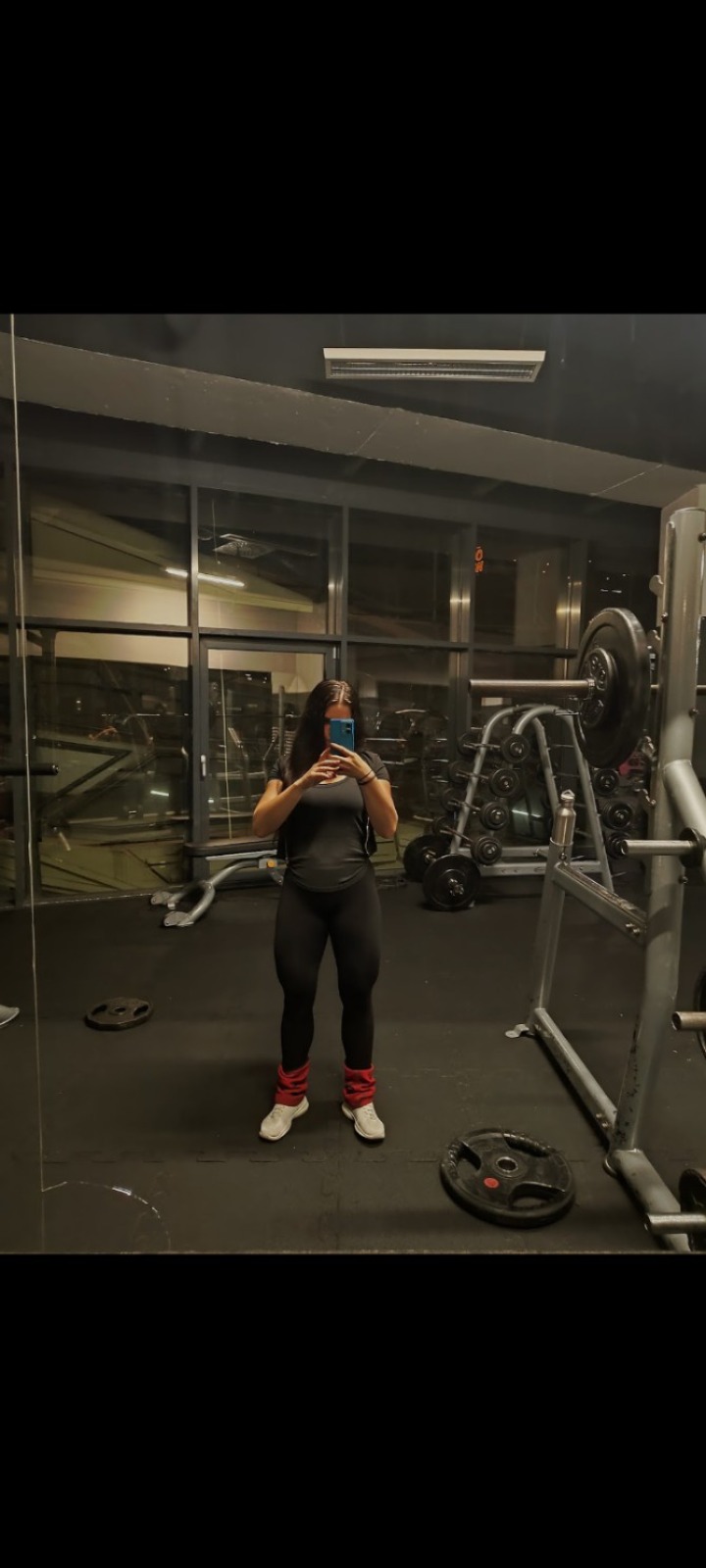 Kobieta w sportowym stroju, z obciążnikami na kostkach, robi selfie w lustrze na tle siłowni z maszynami i sztangami. Widoczne obciążenia na podłodze.