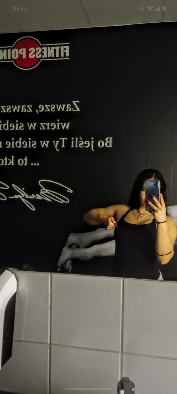 Kobieta robi selfie w lustrze na tle ściany z napisem 'Fitness Point'. Widoczne napięte mięśnie ramion. Ciemne włosy, czarny top i czerwone paznokcie.