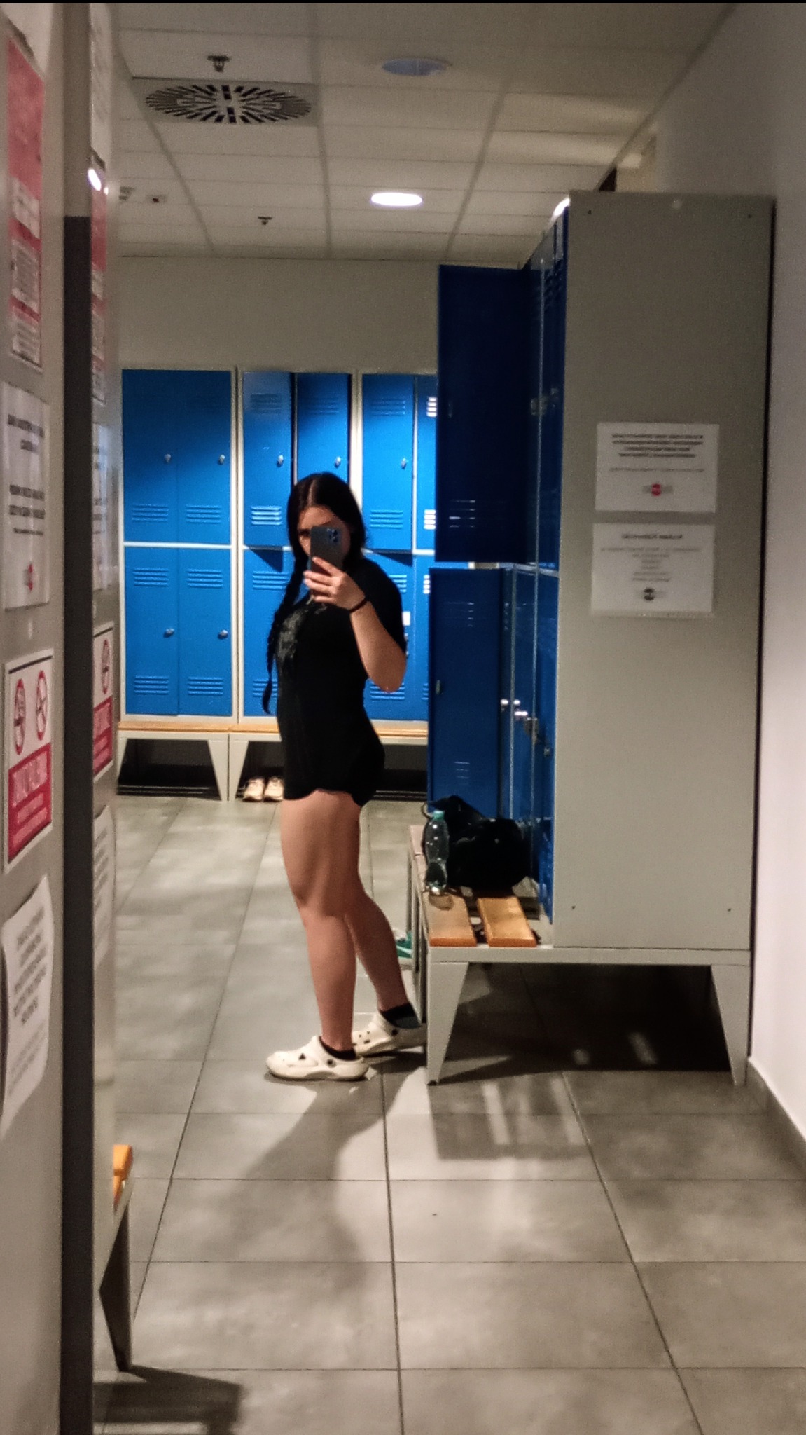 Osoba robi selfie w szatni siłowni na tle niebieskich szafek. Widoczna butelka wody i torba sportowa na ławce. Ubrana w czarny strój sportowy i białe obuwie.