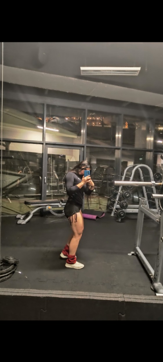 Kobieta w sportowym stroju robi selfie w lustrze na tle siłowni. Na nogach obciążniki, w tle sprzęt do ćwiczeń i okna z widokiem na miasto.