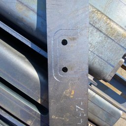 Krysteel - Metalowy element z wyciętym kształtem litery 'U' z dwoma otworami, leżący na tle metalowych profili. Widoczne ślady obróbki.