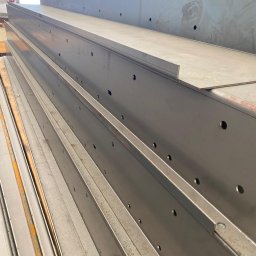 Krysteel - Stalowe belki z otworami ułożone warstwami, widoczne ślady obróbki. Szare, metaliczne wykończenie. Elementy konstrukcyjne w hali.