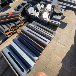 Krysteel - Stalowe elementy konstrukcyjne: profile zamknięte, płaskowniki z otworami i elementy z wyciętymi uśmiechami, ułożone na palecie. Detal obróbki metalu.