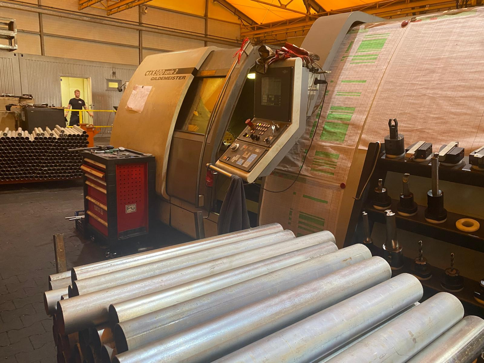 Obrabiarka CNC Gildemeister CTX 500 Serie 2 z narzędziami skrawającymi i stosem metalowych rur. W tle widoczny pracownik w hali.