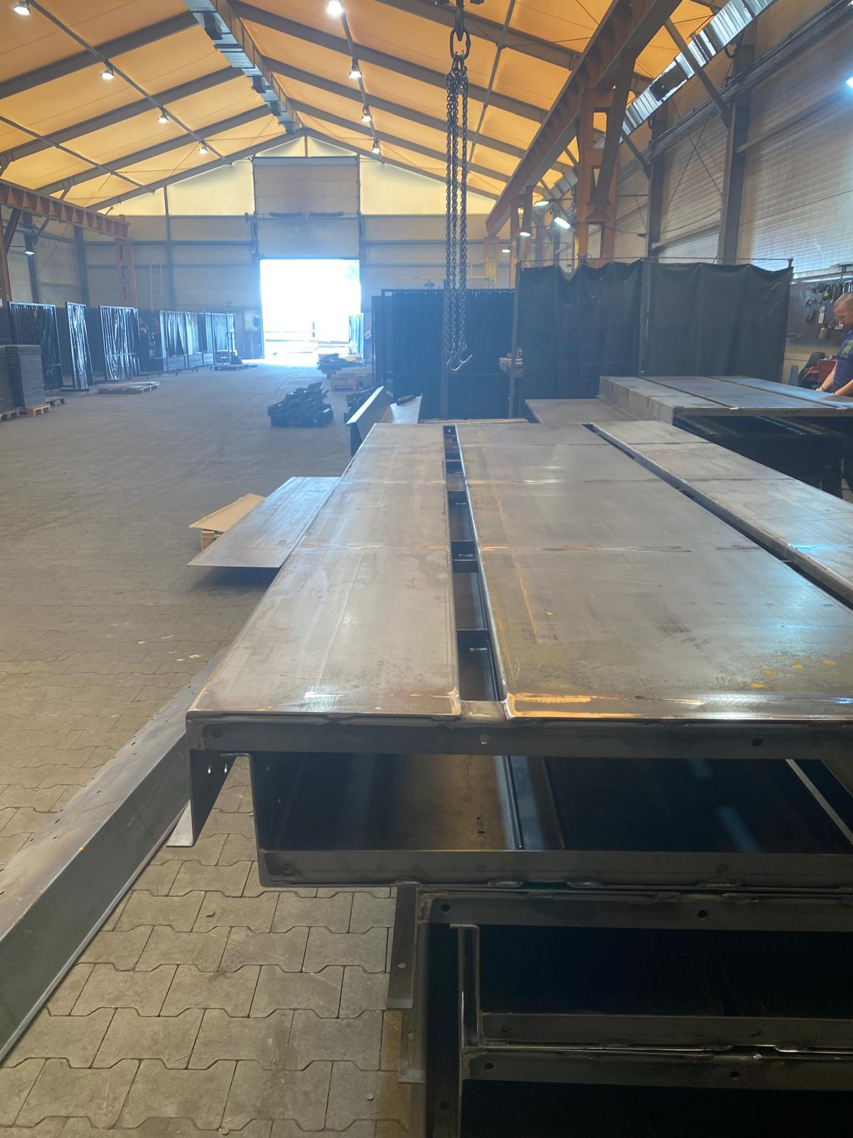 Wnętrze hali produkcyjnej z elementami konstrukcji stalowej w trakcie montażu, podwieszonymi na łańcuchu. Widoczna suwnica i materiały składowane w tle.