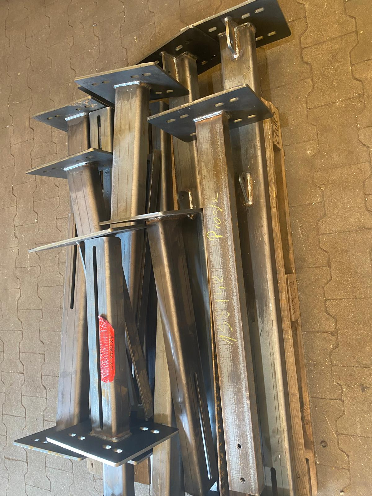 Stalowe słupy z przyspawanymi podstawami, ułożone na podłodze z kostki. Widoczne spawane łączenia i żółte oznaczenia na jednym z elementów.