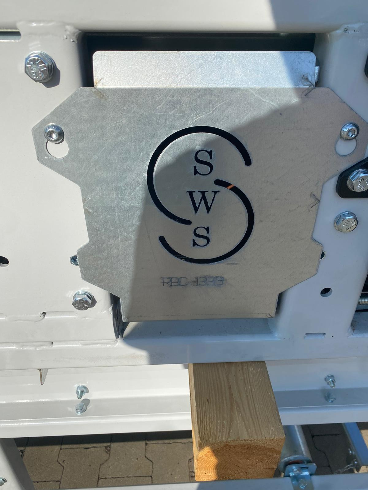 Metalowa płyta z logo SWS przymocowana śrubami do konstrukcji, wsparta na drewnianym elemencie. Widoczne detale spawania i wykończenia.