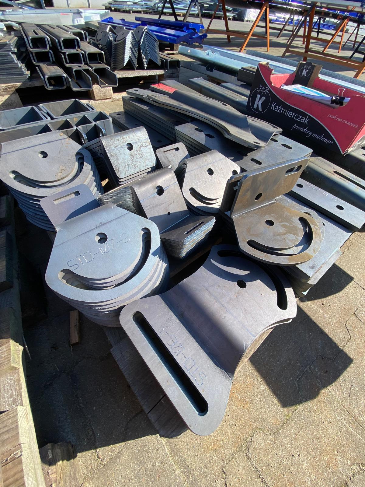 Stalowe elementy konstrukcyjne z otworami i wycięciami, ułożone na palecie. Widoczne profile zamknięte i kątowniki. Na pierwszym planie detale z oznaczeniami STD-107-E.