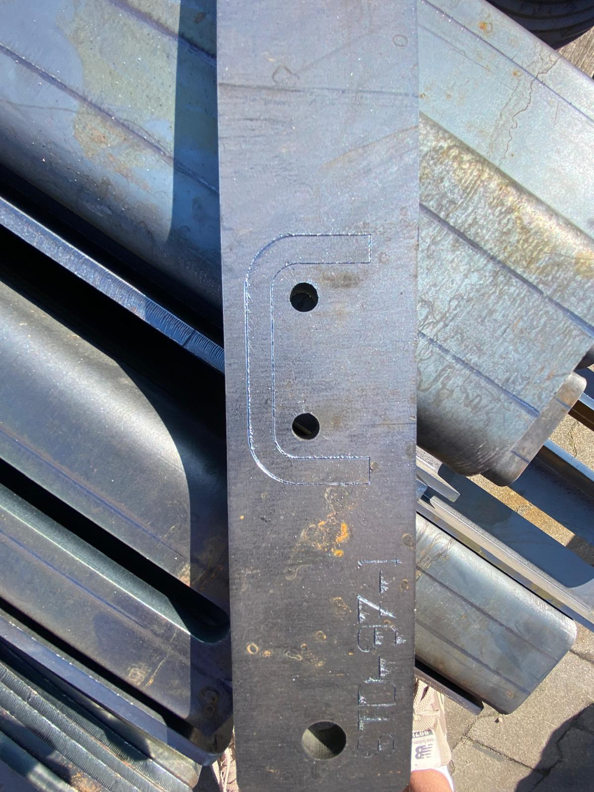 Metalowy element z wyciętym kształtem litery 'U' z dwoma otworami, leżący na tle metalowych profili. Widoczne ślady obróbki.