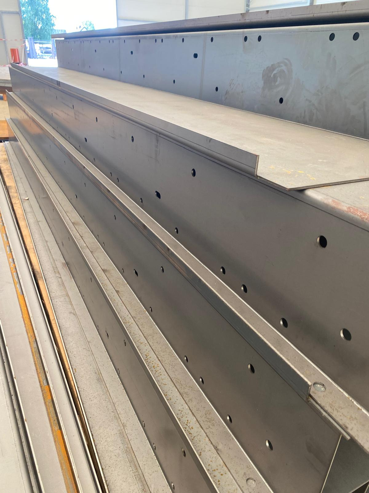 Stalowe belki z otworami ułożone warstwami, widoczne ślady obróbki. Szare, metaliczne wykończenie. Elementy konstrukcyjne w hali.