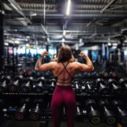 Kobieta prezentuje umięśnione plecy i ramiona w siłowni, na tle regałów z hantlami. Widoczny tatuaż na ramieniu i strój sportowy z odkrytymi plecami.