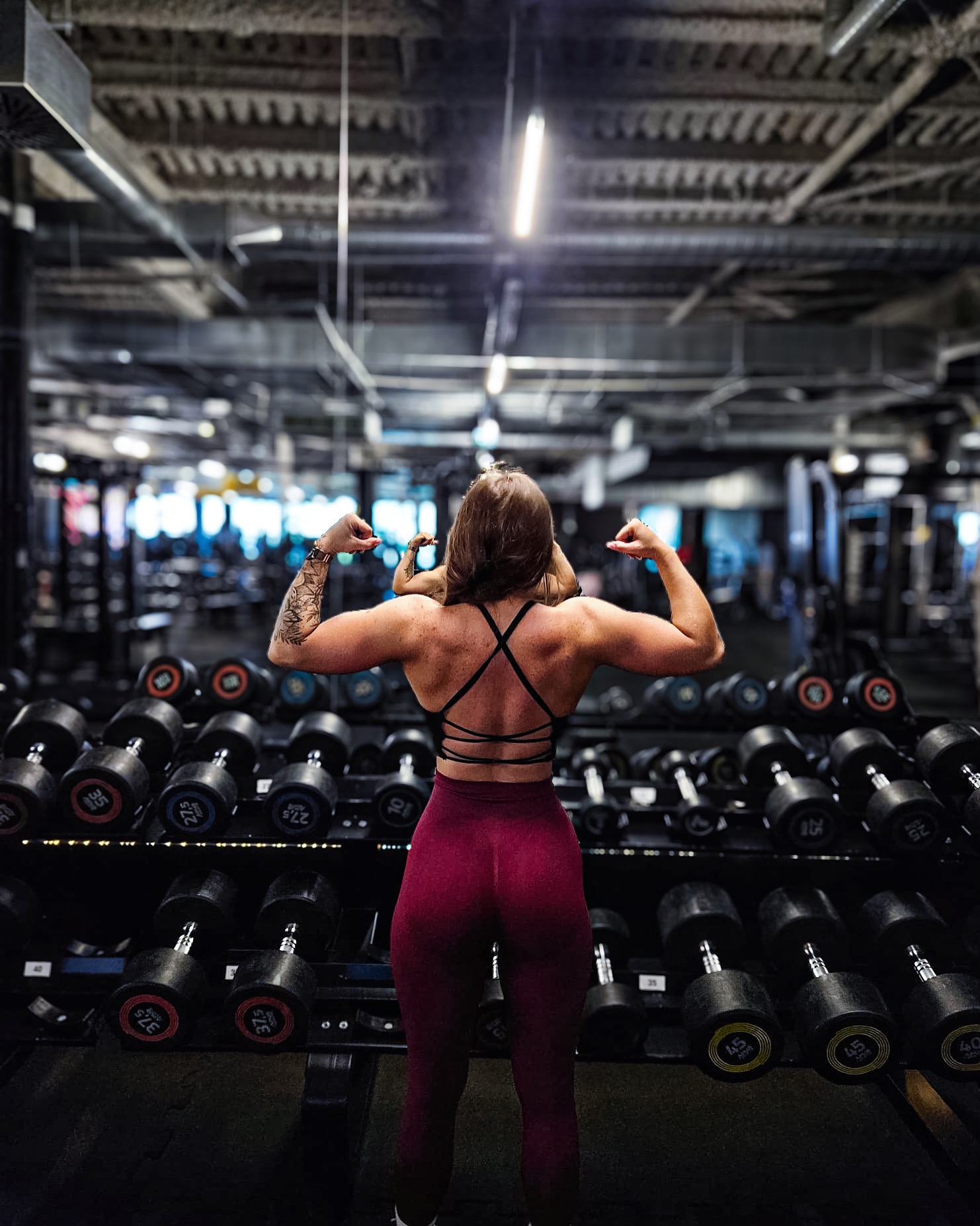 Kobieta prezentuje umięśnione plecy i ramiona w siłowni, na tle regałów z hantlami. Widoczny tatuaż na ramieniu i strój sportowy z odkrytymi plecami.