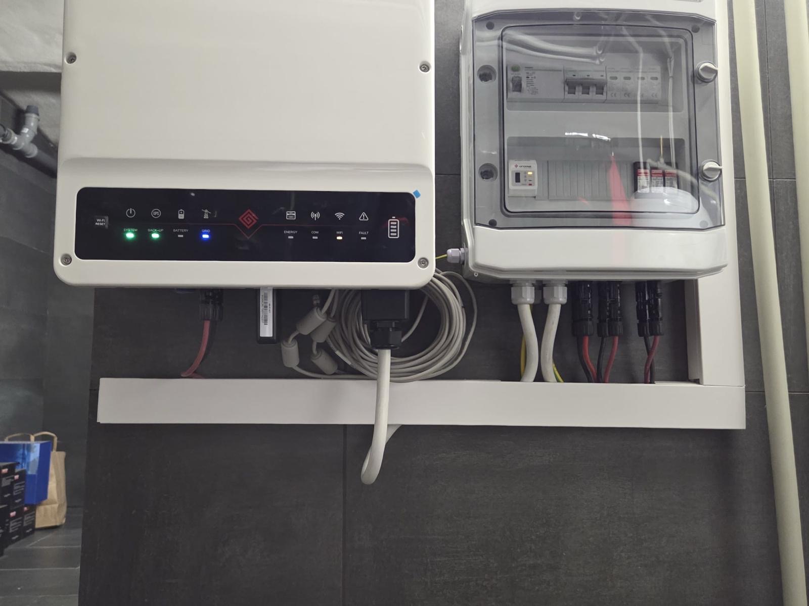 Biała skrzynka elektryczna z widocznymi bezpiecznikami i przewodami, zamontowana na szarej ścianie. Obok falownik z diodami LED i kablem. Detal instalacji.