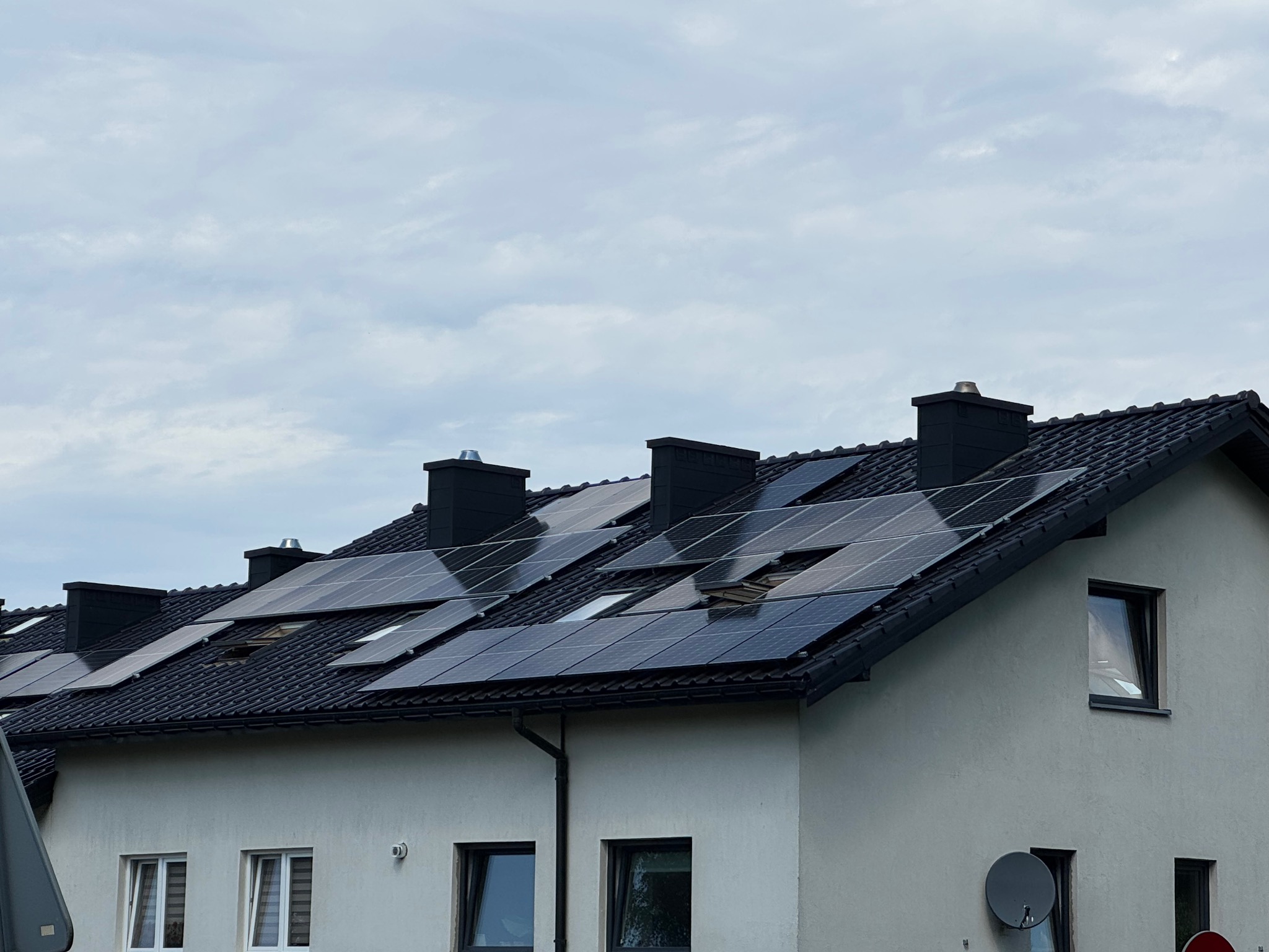 Panele słoneczne na dachu domu jednorodzinnego z czarną dachówką i kominami. Widok z dołu na pochmurne niebo, antena satelitarna na ścianie budynku.