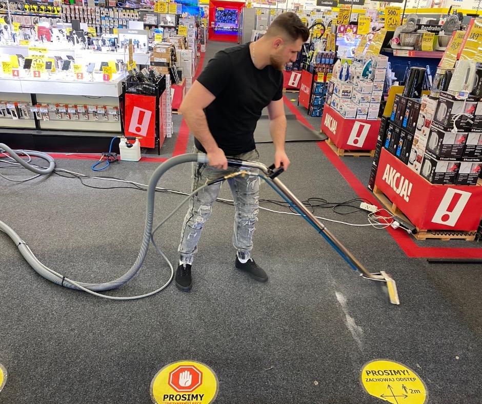 Mężczyzna czyści dywan w sklepie za pomocą profesjonalnego sprzętu. Widoczne żółte naklejki ostrzegawcze na podłodze i regały z towarem w tle.