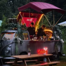 Rustykalny bar mobilny w stylu vintage, umieszczony na zewnątrz, z neonowym napisem 'Drinkey', obsługiwany przez dwóch barmanów.