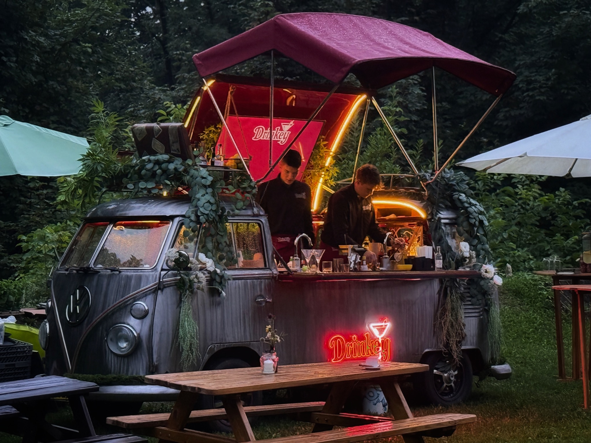 Rustykalny bar mobilny w stylu vintage, umieszczony na zewnątrz, z neonowym napisem 'Drinkey', obsługiwany przez dwóch barmanów.