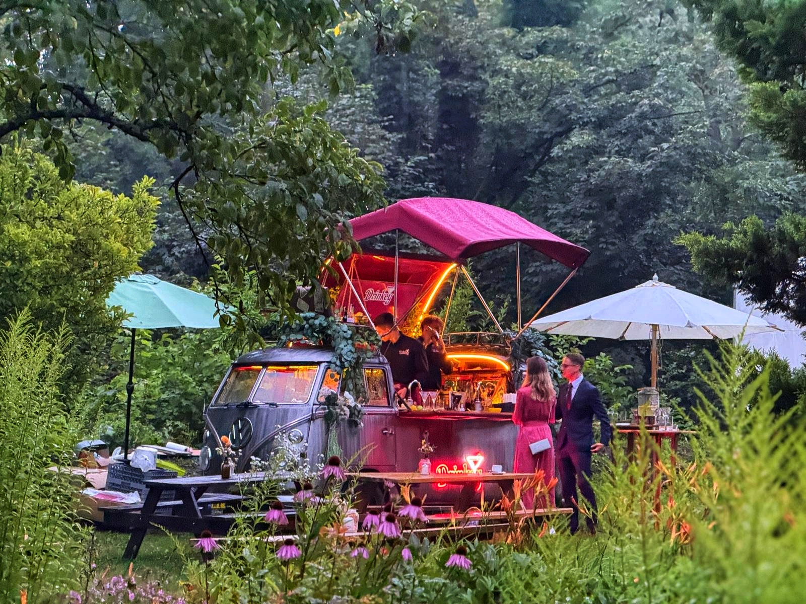 Rustykalny bar mobilny w stylu retro, umieszczony w ogrodzie, obsługiwany przez barmana, z klientami. Całość oświetlona dekoracyjnym neonem.