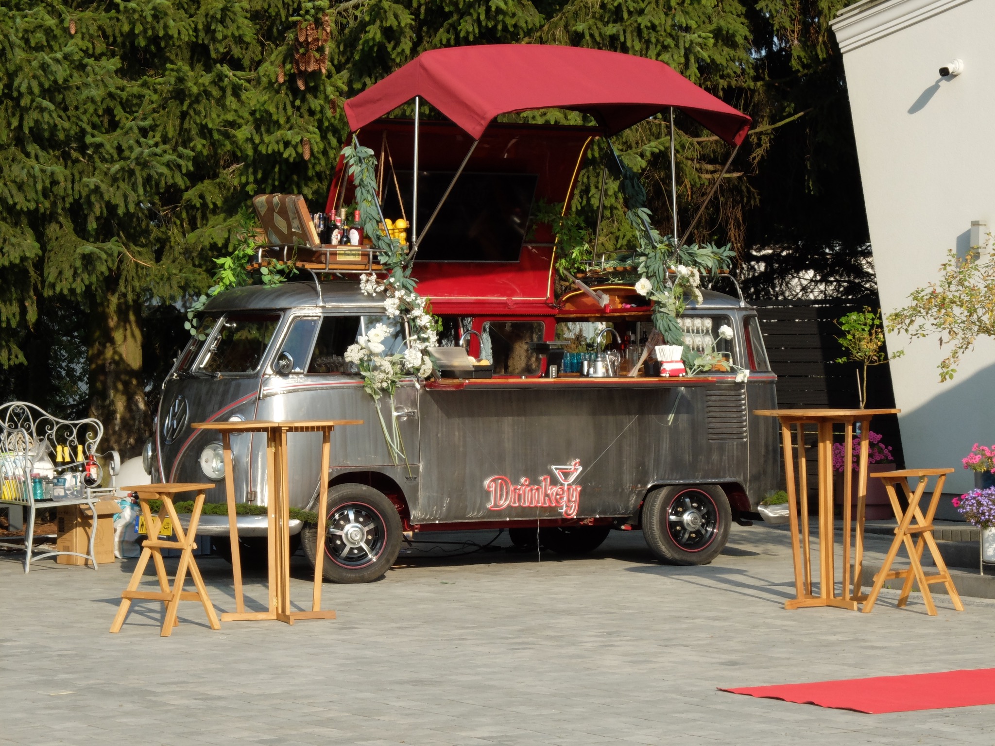 Rustykalny bar mobilny 'Drinkey' w stylu vintage VW bus, udekorowany kwiatami, z czerwonym baldachimem, ustawiony na zewnątrz z drewnianymi stolikami.