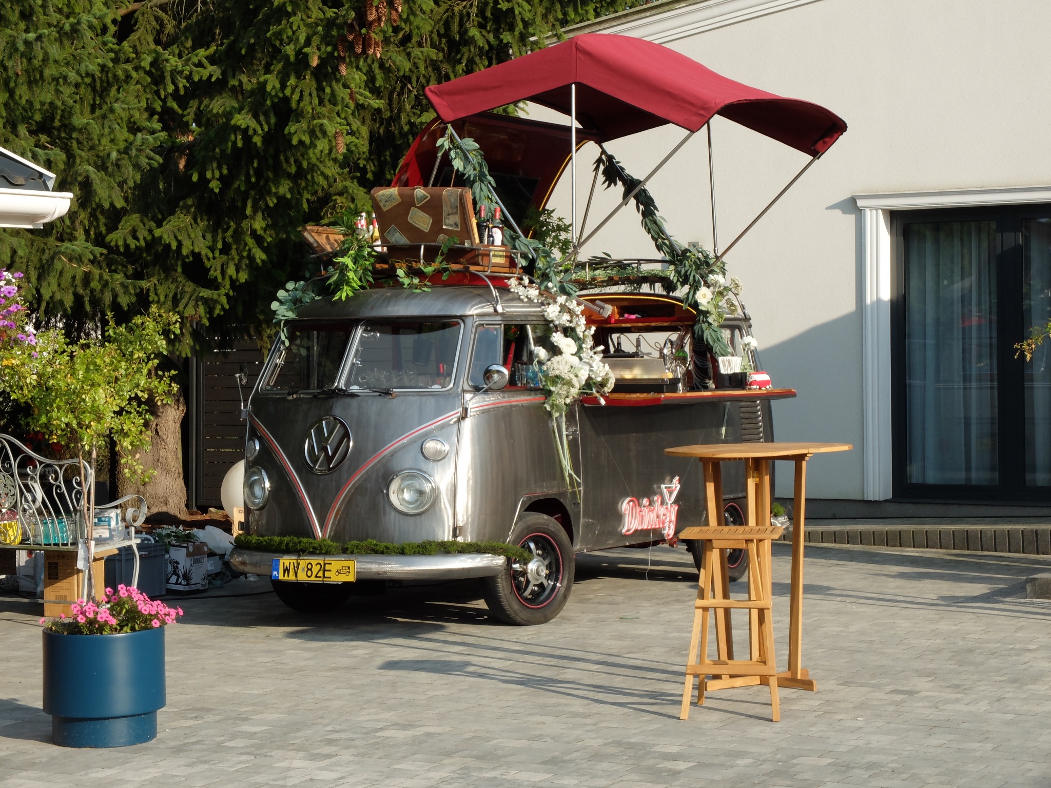 Stylizowany food truck VW Bus z dekoracjami roślinnymi i drewnianym stolikiem barowym, ustawiony na zewnątrz budynku, idealny na imprezy plenerowe.