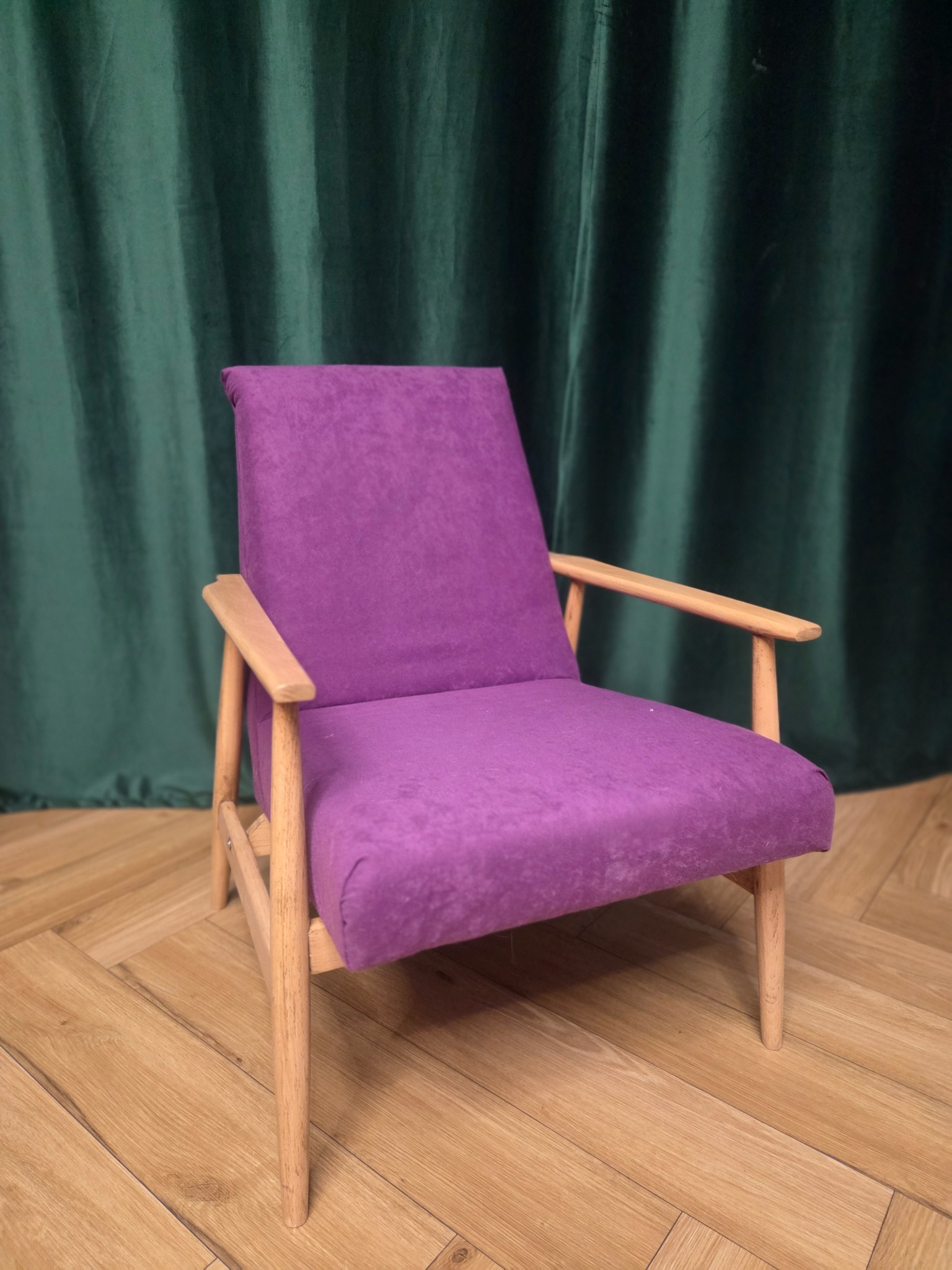 Fioletowy fotel z drewnianymi nogami na parkiecie, na tle zielonej zasłony. Ujęcie z bliska, prezentujące detale tapicerki i konstrukcji mebla.