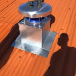 Błyszczący, metalowy kominek wentylacyjny na pomarańczowym dachu z blachy falistej. Na kominku leży wiertarka. W tle widoczny cień osoby.