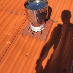 Varsobud POLSKA - Pomarańczowy dach z blachy trapezowej z kominem wentylacyjnym i cieniem osoby. Widoczne ślady uszczelniania wokół podstawy komina.