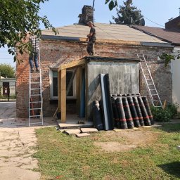 Varsobud POLSKA - Dwóch mężczyzn na drabinach i dachu podczas prac dekarskich przy budynku z cegły. Widoczne rolki papy i narzędzia.