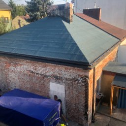 Varsobud POLSKA - Widok z góry na budynek z nowym, ciemnym pokryciem dachowym i ceglaną elewacją. Obok zaparkowane auto dostawcze pod plandeką. W tle zabudowa miejska.