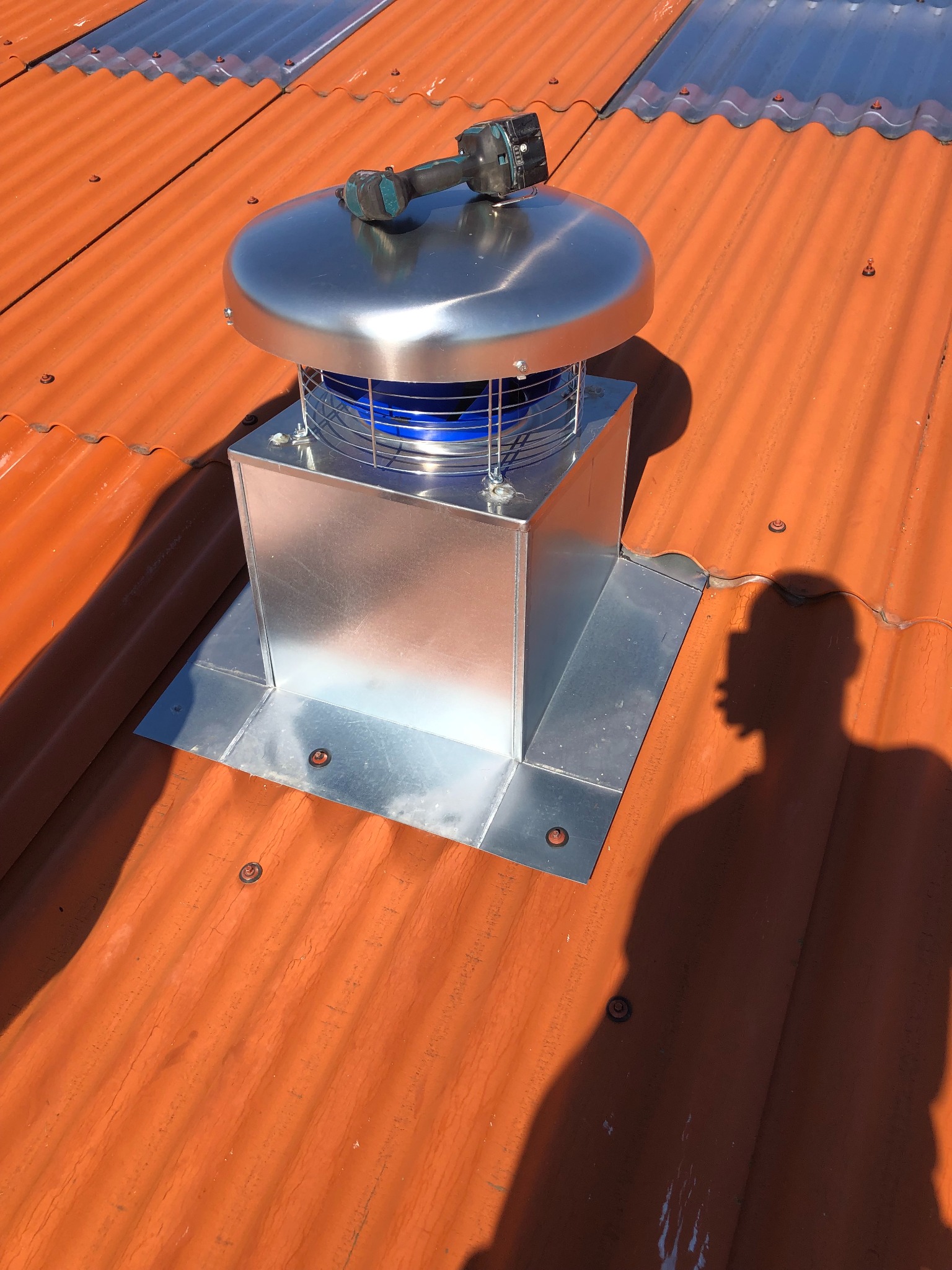 Błyszczący, metalowy kominek wentylacyjny na pomarańczowym dachu z blachy falistej. Na kominku leży wiertarka. W tle widoczny cień osoby.