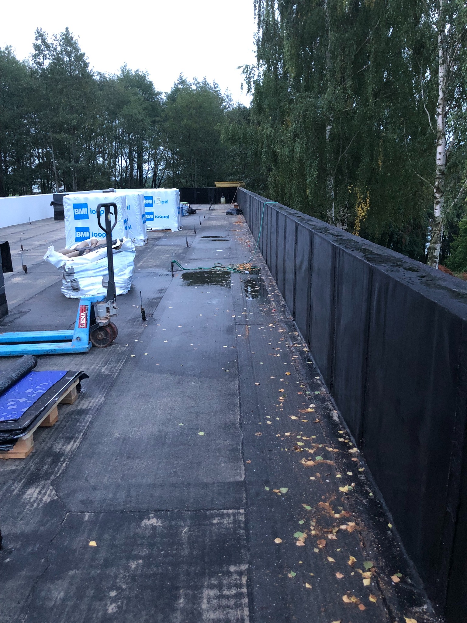 Płaski dach pokryty papą z paletą materiałów budowlanych i wózkiem paletowym. Widoczne obróbki blacharskie i jesienne liście na powierzchni.