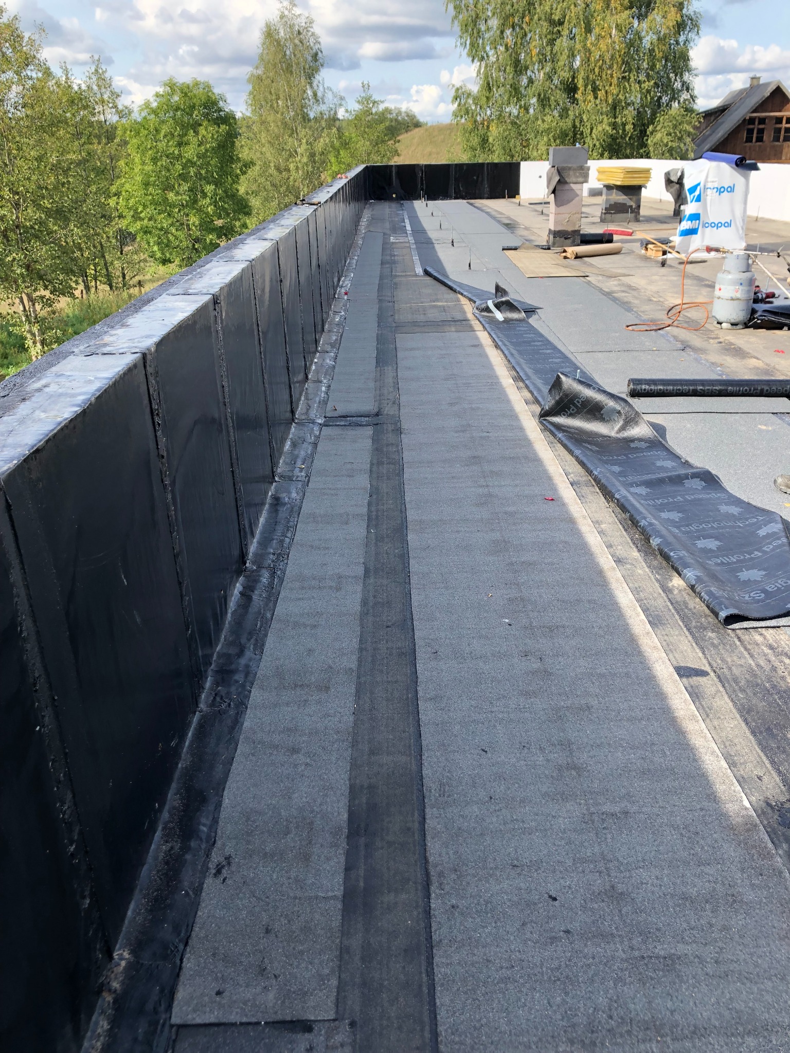 Płaski dach pokryty papą termozgrzewalną, częściowo ułożoną. Widoczne narzędzia i materiały dekarskie na tle zieleni drzew i błękitnego nieba.