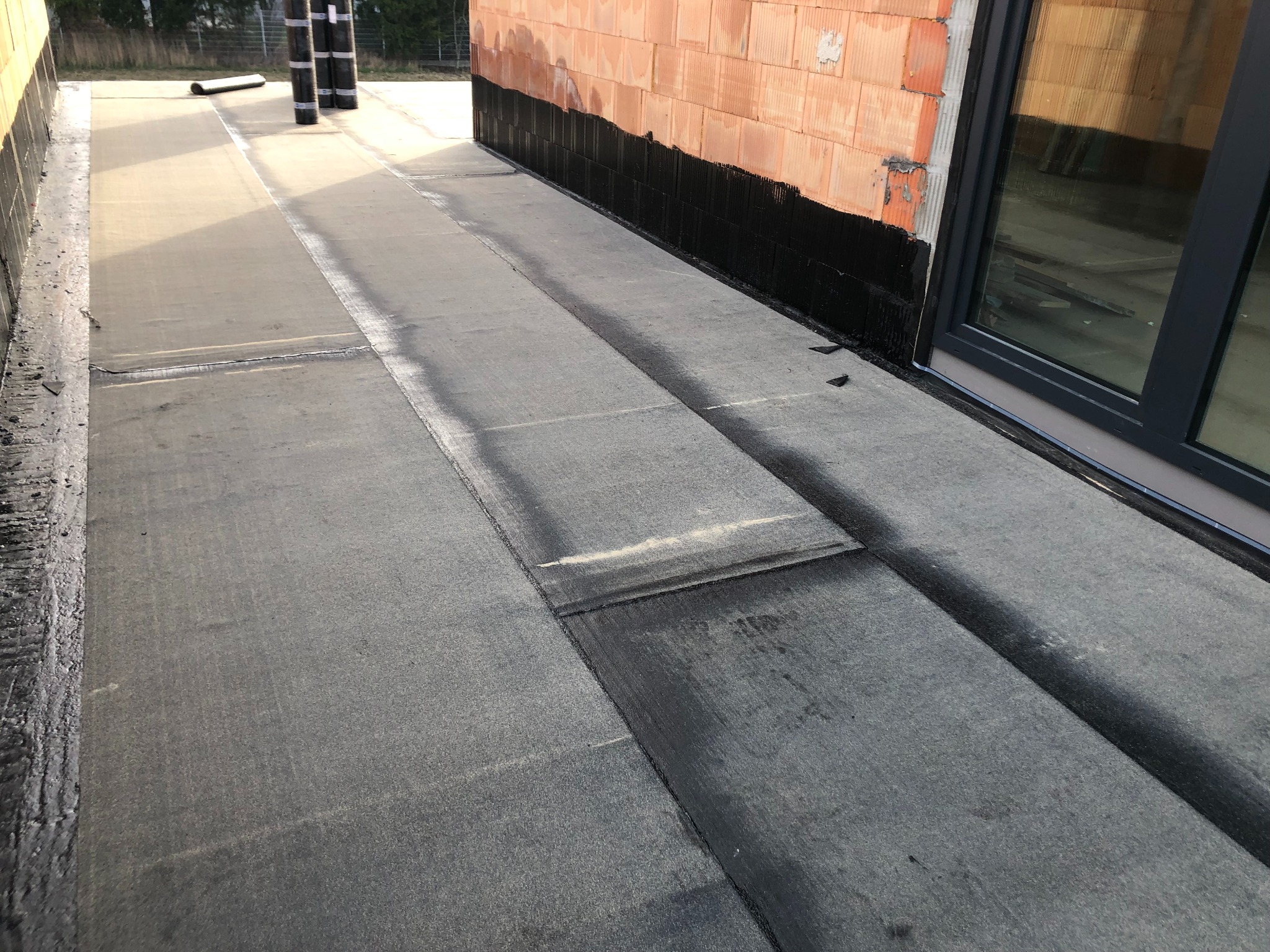 Płaski dach pokryty papą termozgrzewalną, widoczne łączenia i fragment ściany z cegły. Na dachu leżą rolki papy. Nowe okno dachowe w tle.