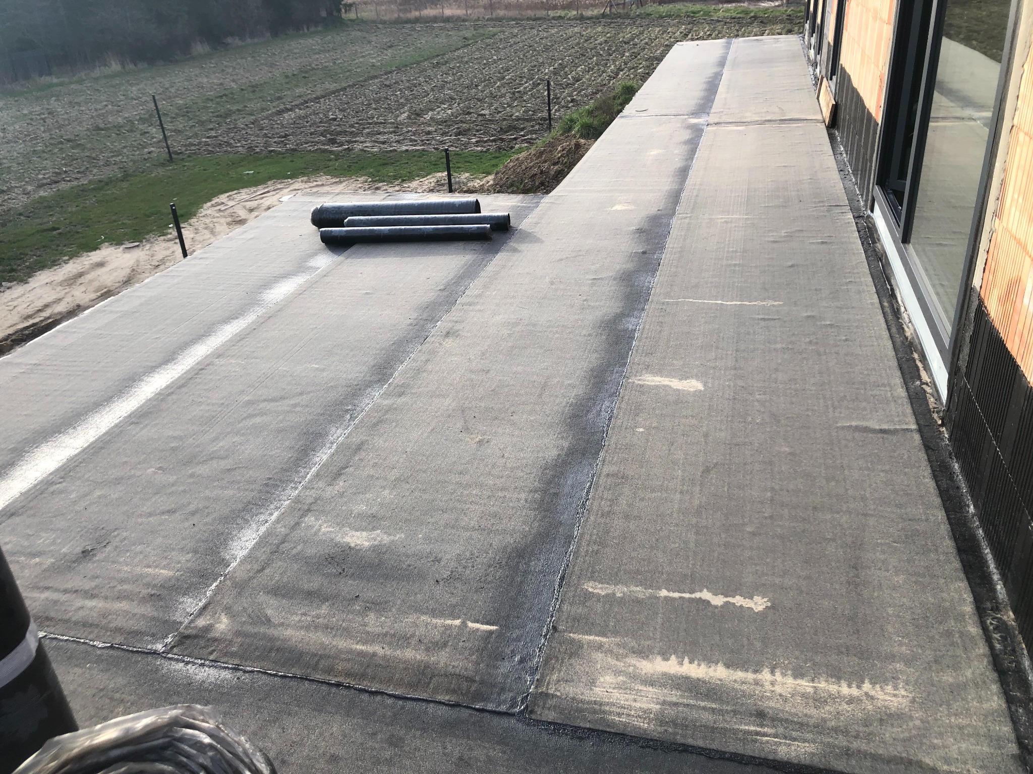 Wykończony, płaski dach pokryty papą, z widocznymi łączeniami. W tle pole i fragment budynku z oknami. Część rolki papy w dolnym rogu kadru.