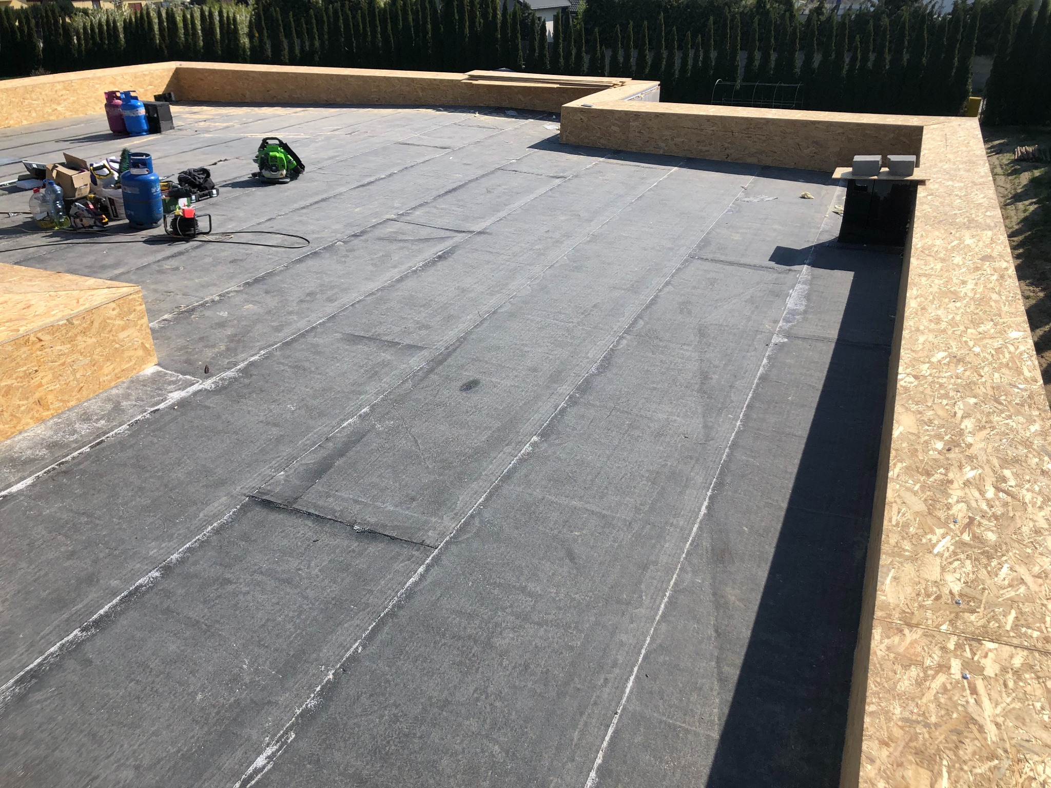 Płaski dach pokryty papą, obłożony drewnianą konstrukcją. Widoczne narzędzia i materiały budowlane. Dach przygotowany do dalszych prac wykończeniowych.