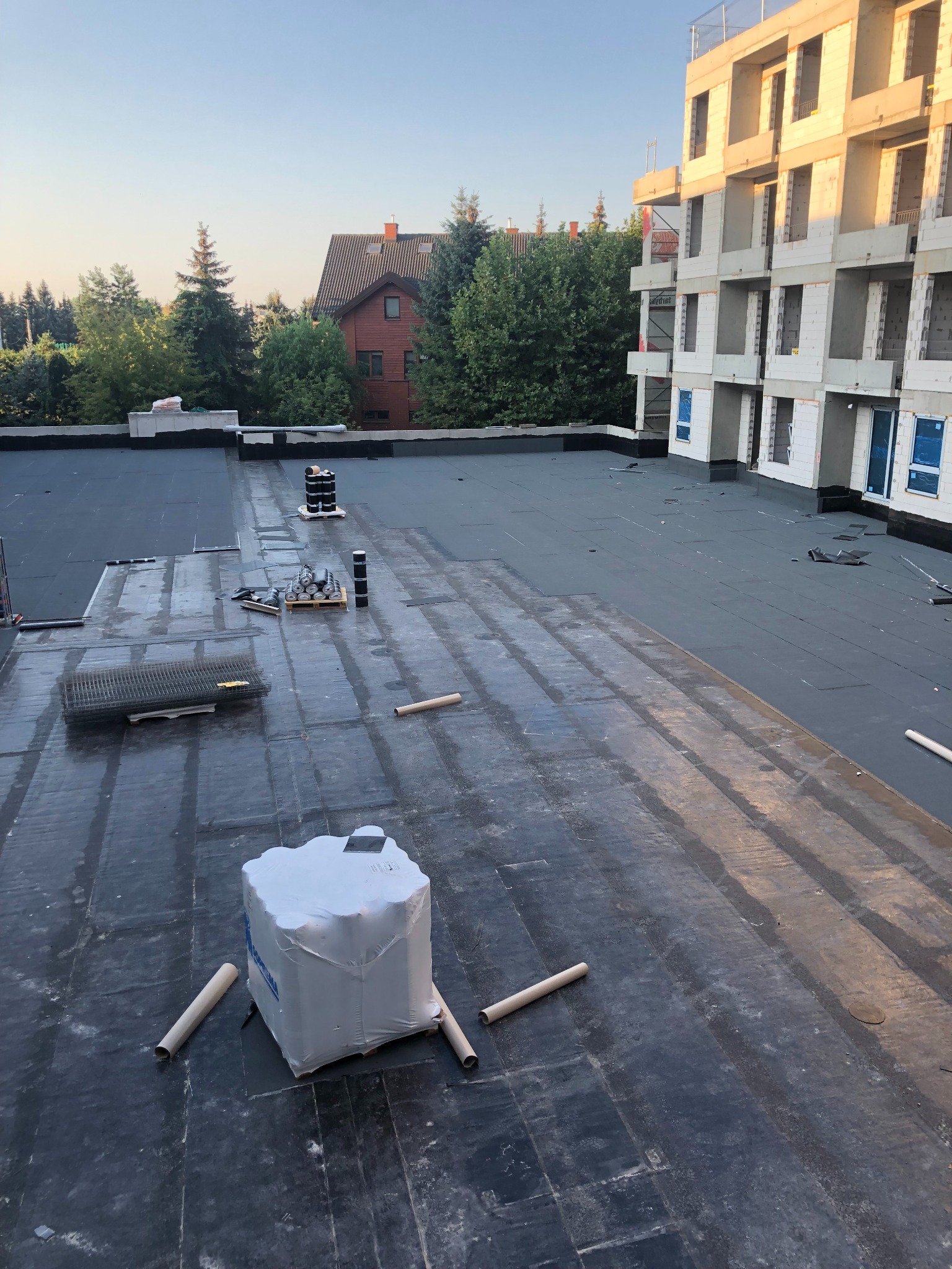 Prace dekarskie na płaskim dachu budynku w budowie. Widoczne materiały izolacyjne, narzędzia i elementy konstrukcyjne. W tle budynek mieszkalny i zieleń.