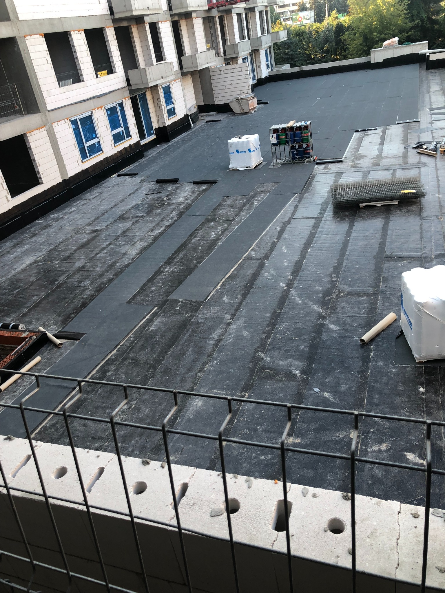 Widok z góry na pokryty czarną papą dach budynku w trakcie budowy. Na dachu materiały budowlane i narzędzia. W tle drzewa i fragmenty innych budynków.