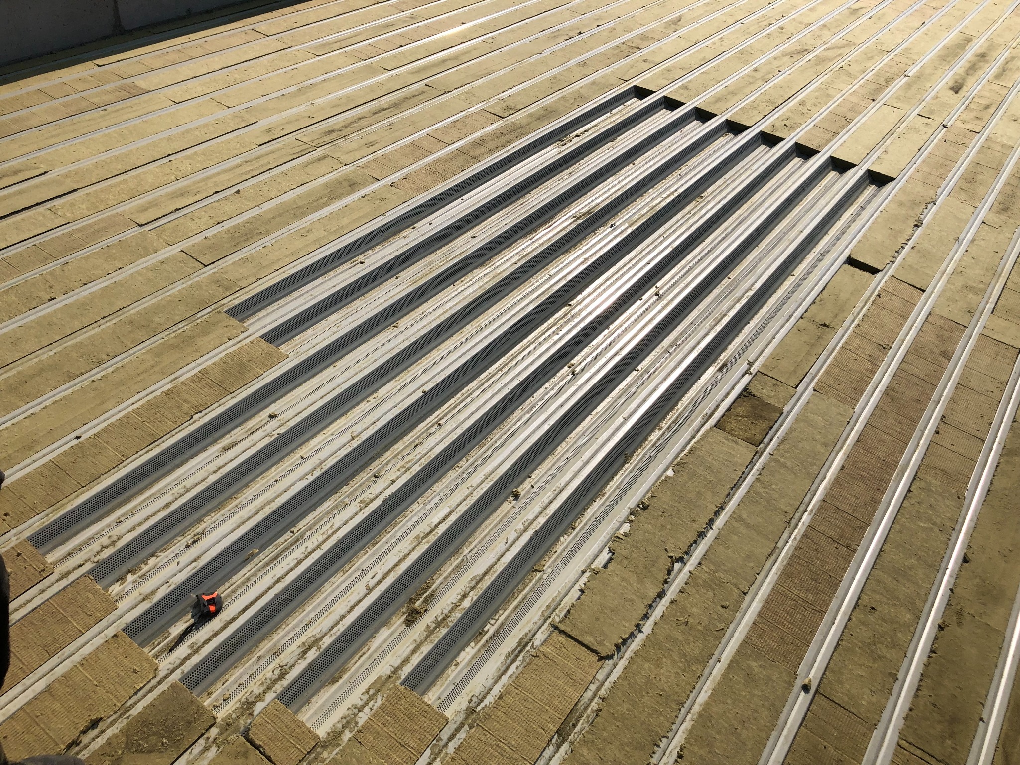 Metalowa konstrukcja dachu z częściowo ułożoną izolacją z wełny mineralnej w beżowym odcieniu. Widoczny fragment dłoni i narzędzie pomiarowe.