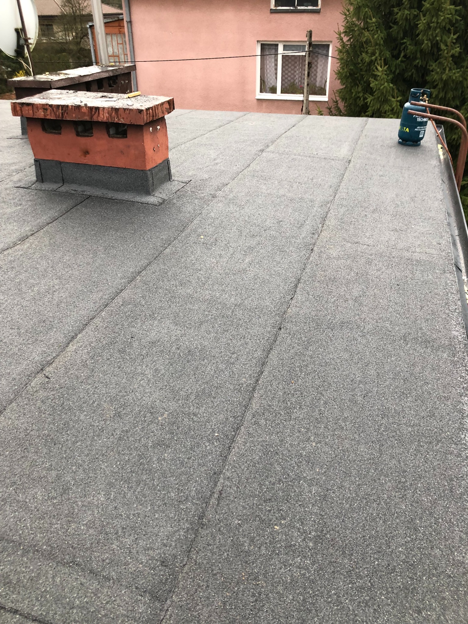 Szary, płaski dach pokryty papą z kominem i butlą gazową w tle. Widoczne łączenia papy. Dom z oknem i zieleń w tle. Ujęcie z góry, podkreślające teksturę pokrycia.
