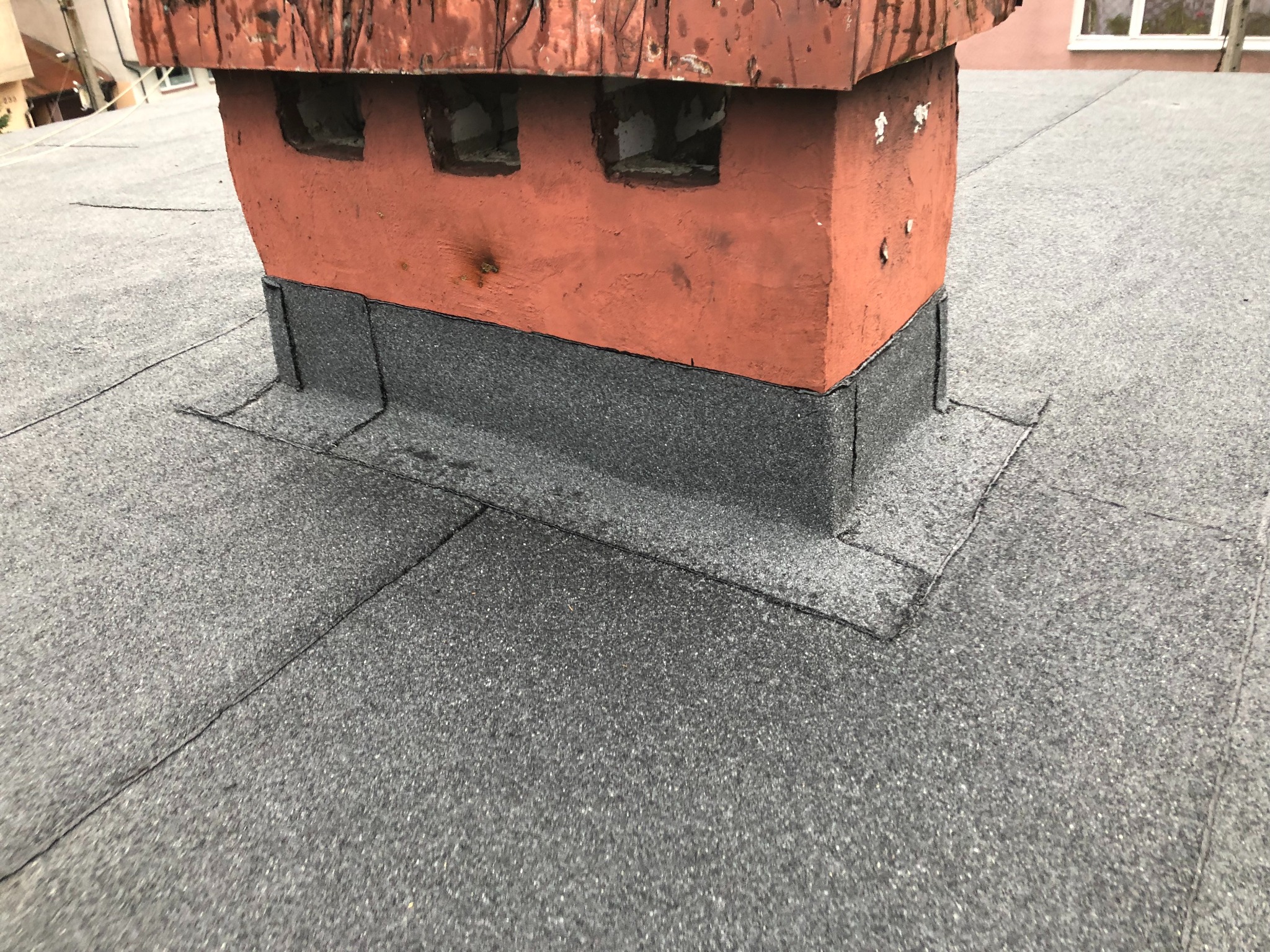 Szary dach z papy z kominem z cegły i obróbką blacharską. Widoczne łączenia papy, detale wykończenia komina. Ujęcie z góry.