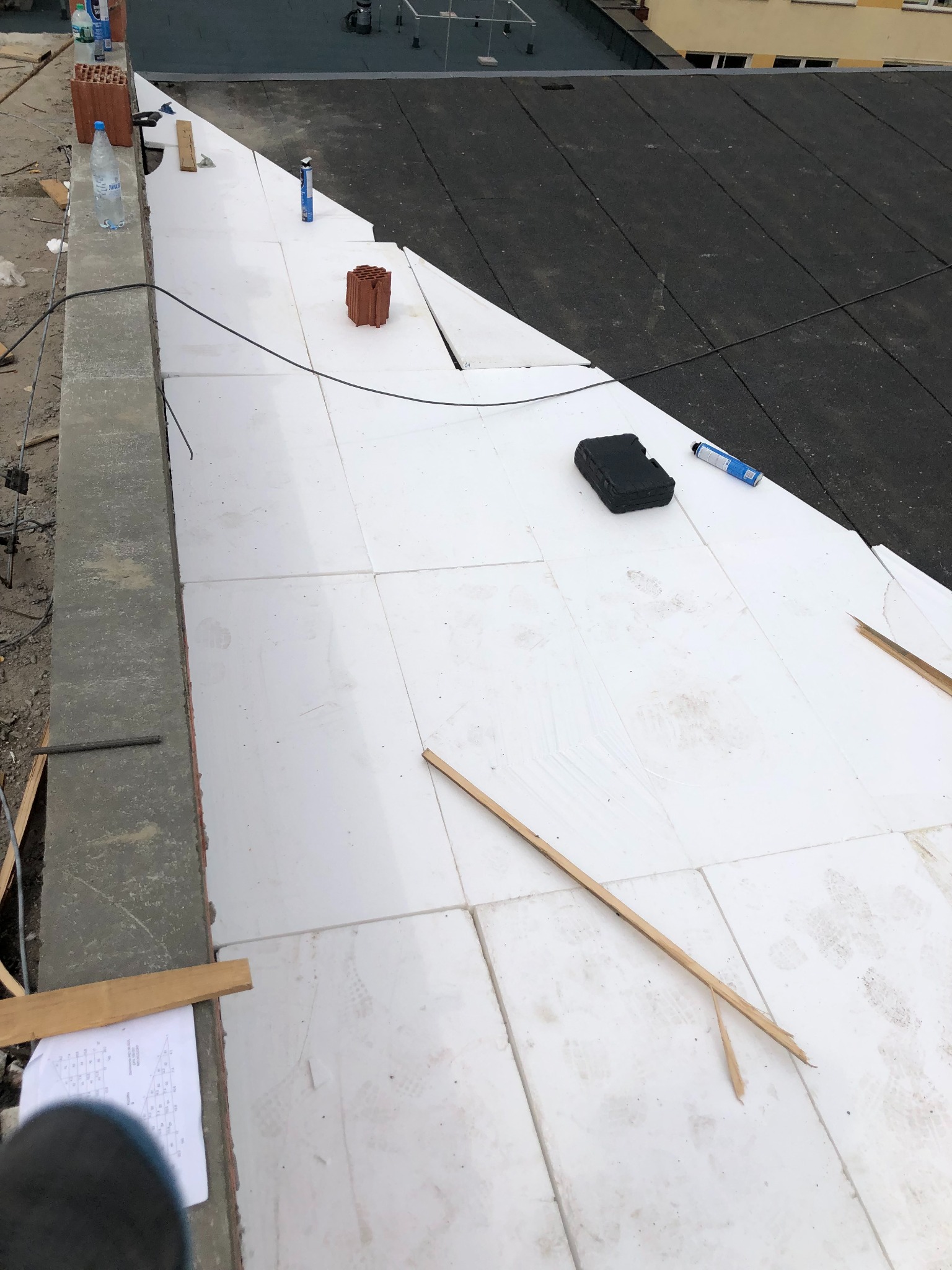 Ocieplenie dachu styropianem w trakcie prac. Widoczne narzędzia, cegły, woda i czarna papa. Częściowo ocieplony dach budynku mieszkalnego.