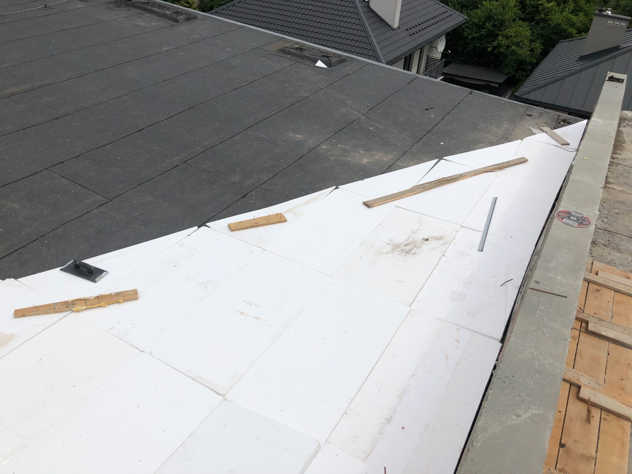 Fragment dachu płaskiego w trakcie ocieplania styropianem, widoczne narzędzia i elementy konstrukcyjne; w tle inne dachy i zieleń.