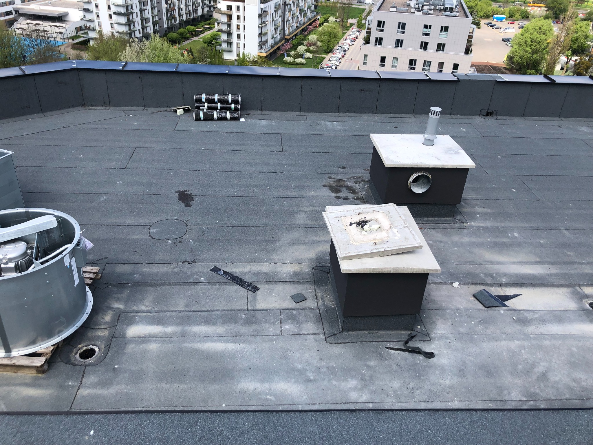 Płaski dach pokryty papą z elementami wentylacyjnymi i fragmentami blachy. Widoczne narzędzia i materiały dekarskie. Tło stanowią budynki mieszkalne.