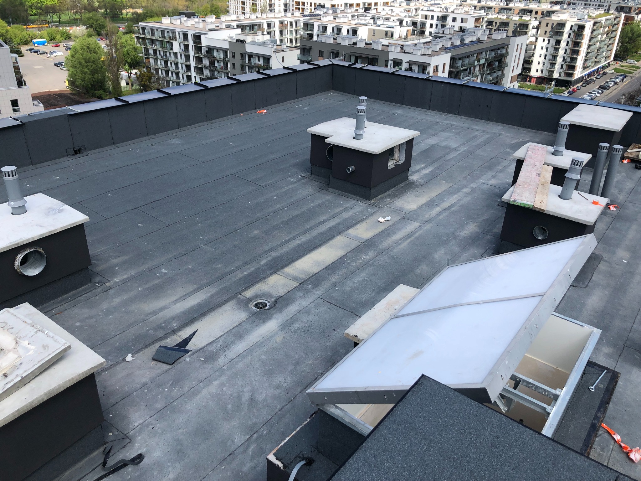 Płaski dach budynku wielorodzinnego z pokryciem bitumicznym, widoczne kominy wentylacyjne i wyłaz dachowy. W tle zabudowa miejska.