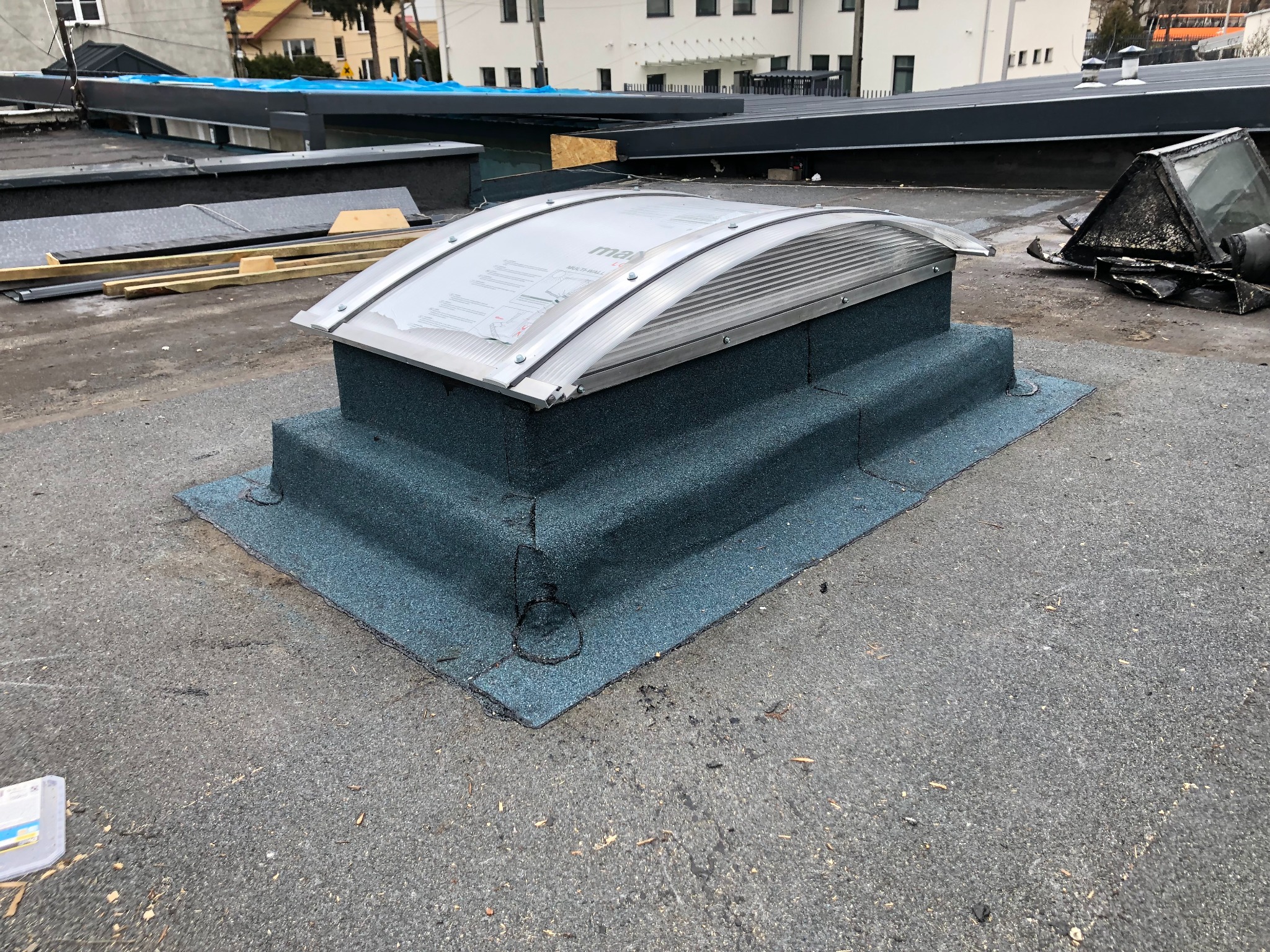 Zamontowany świetlik dachowy na płaskim dachu pokrytym papą, z widocznymi obróbkami blacharskimi i elementami montażowymi. W tle inne elementy dachu i budynki.