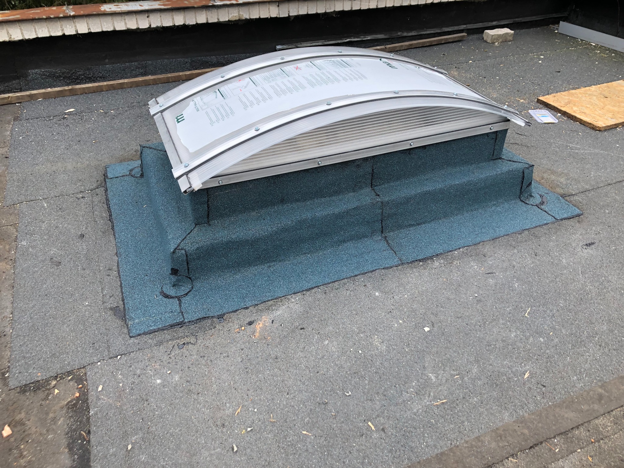 Zamontowany świetlik dachowy na płaskim dachu pokrytym papą. Widoczne detale obróbki blacharskiej wokół świetlika i mocowania.