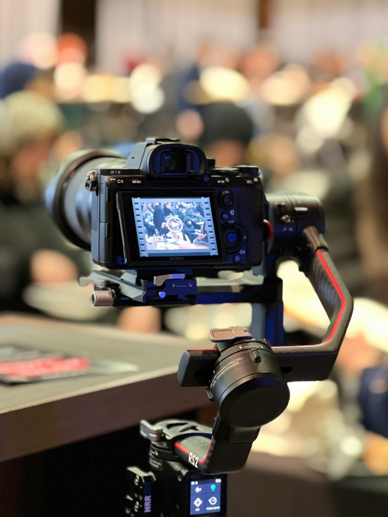 Kamera Sony Alpha 7 III na stabilizatorze DJI RS2, ekran pokazuje nagranie z imprezy firmowej. W tle rozmyte sylwetki uczestników.