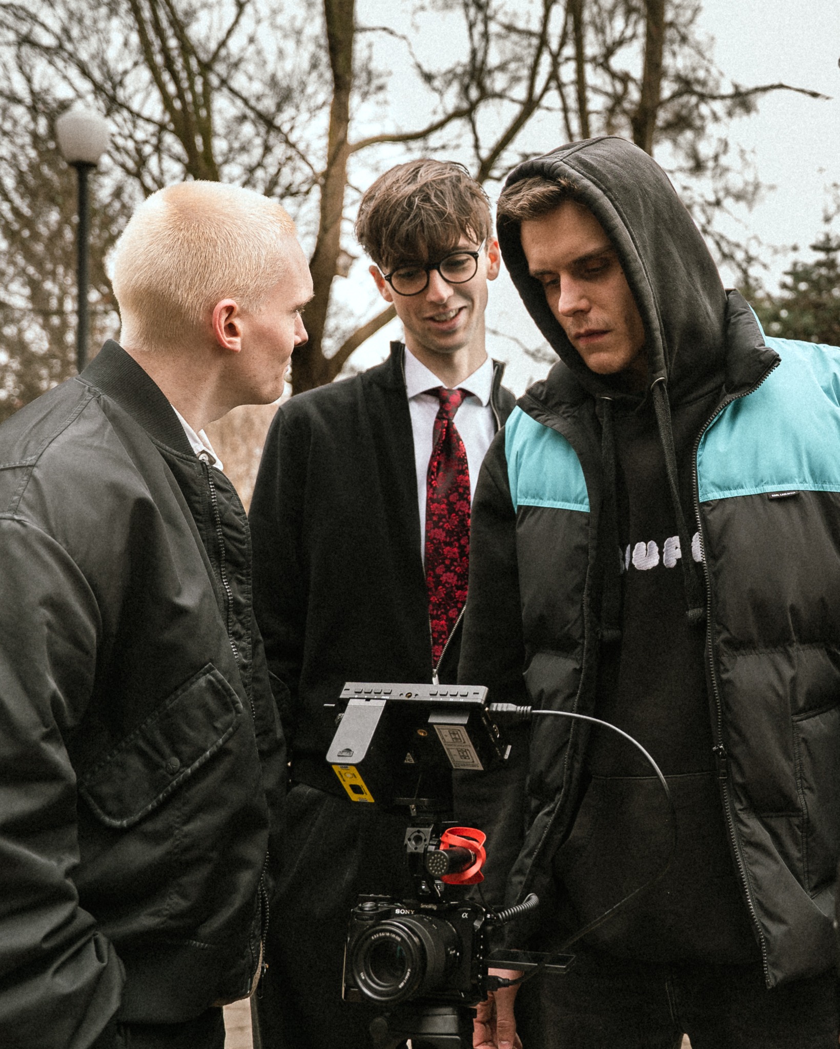 Ekipa filmowa w plenerze: trzech mężczyzn z kamerą Sony na statywie, monitor podglądowy. Jeden w garniturze z czerwoną koszulą, drugi w bluzie z kapturem, trzeci w kurtce.