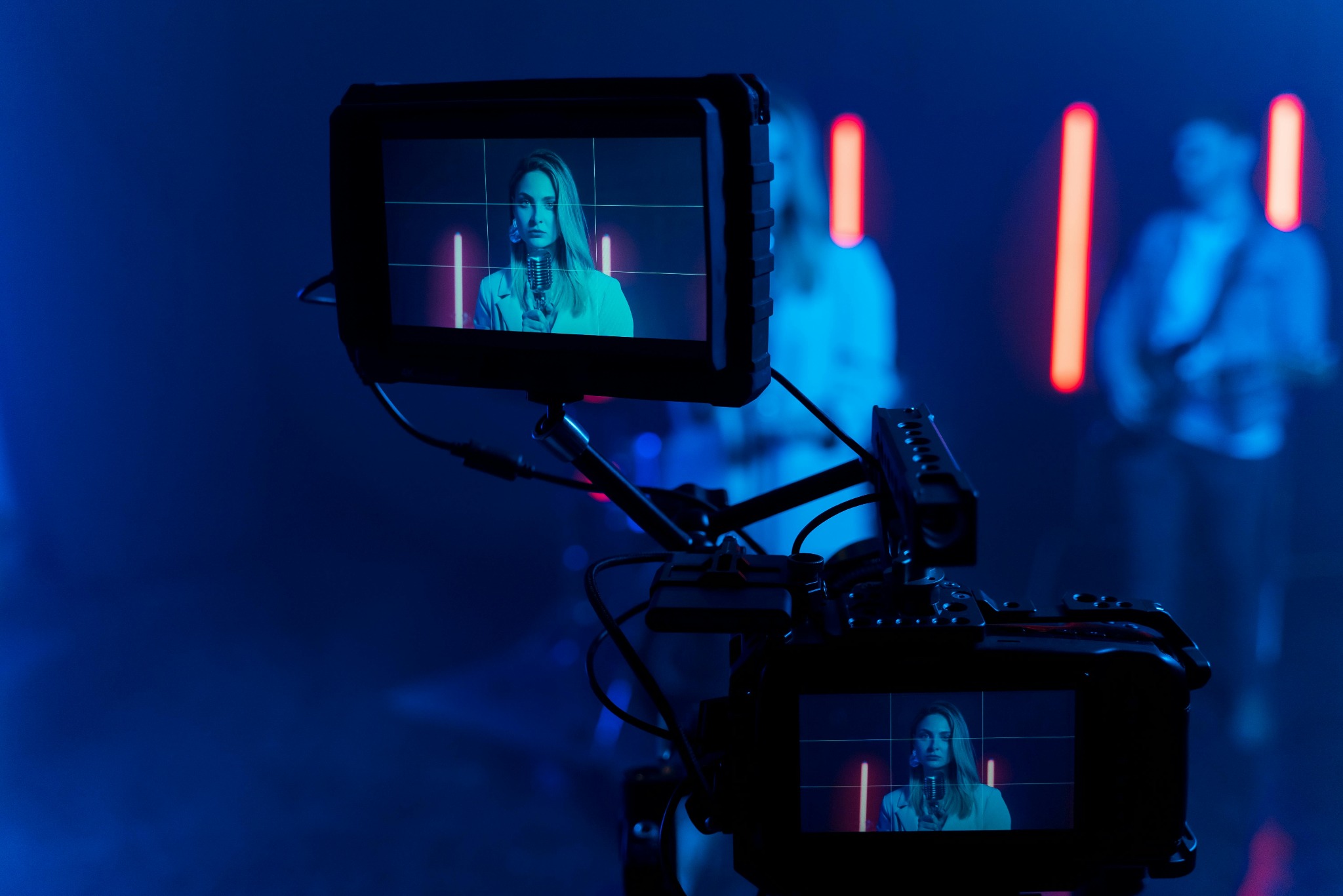 Kamera filmowa rejestruje scenę z wokalistką i muzykami w tle, oświetloną neonowymi lampami. Ekran kamery pokazuje zbliżenie na wokalistkę z mikrofonem.